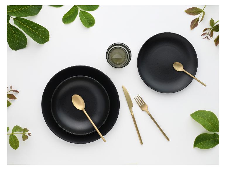 Schwarzes Geschirr mit goldenem Besteck und grünen Blättern auf weißem Hintergrund.