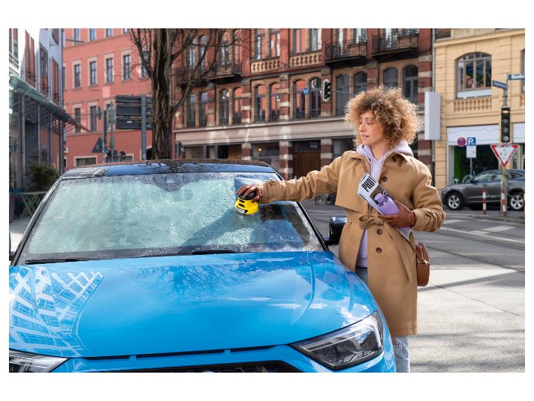 Frau reinigt Autoscheibe mit Eiskratzer, hält Magazin und Wasserflasche.