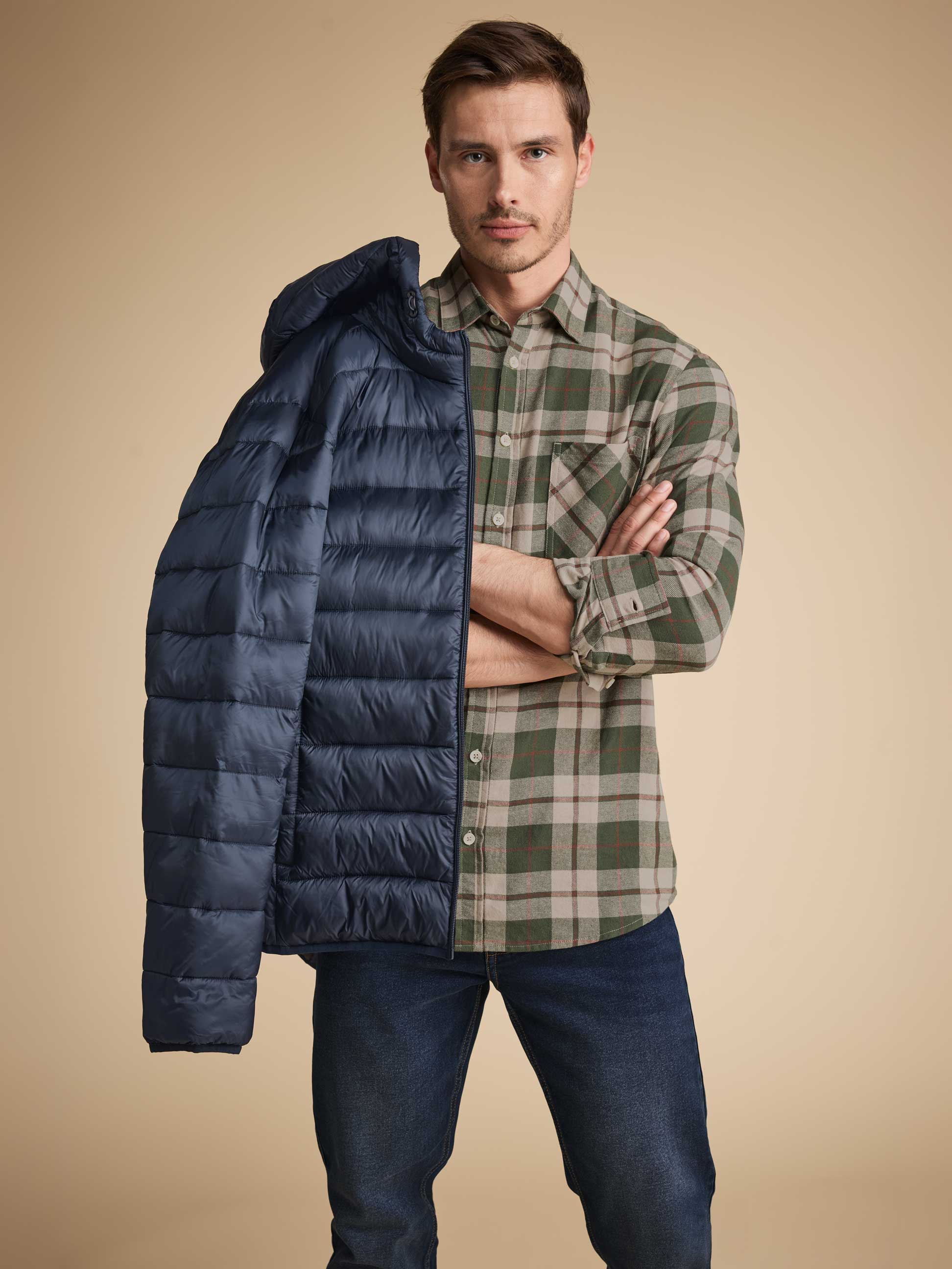 Mann in grünem Karohemd und Jeans, der eine blaue Steppjacke über der Schulter hält.