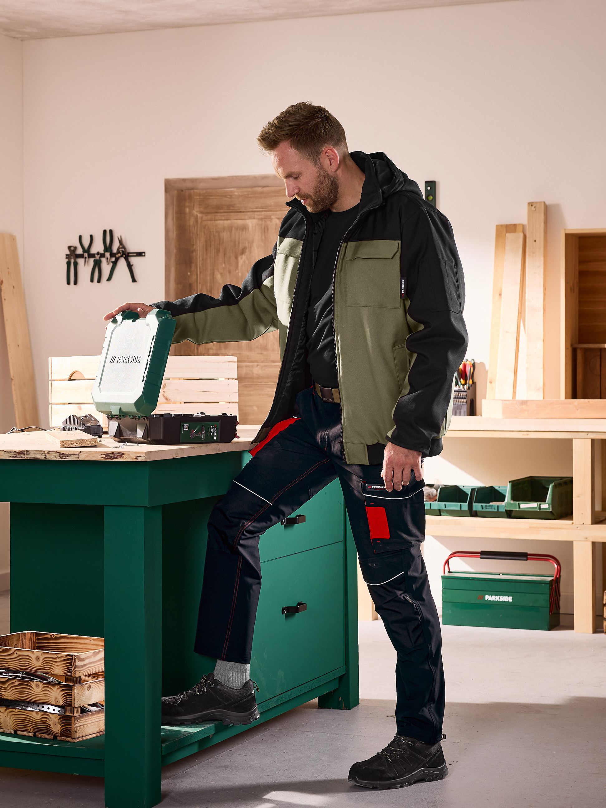 Mann in Arbeitsjacke und -hose öffnet einen Werkzeugkoffer auf einer Werkbank in einer Werkstatt.