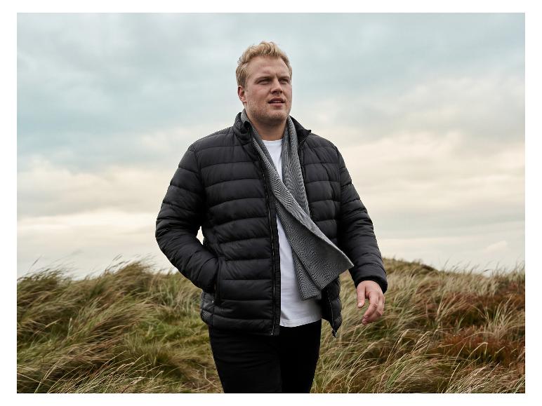 Mann in schwarzer Steppjacke und grauem Schal steht in windiger Landschaft.