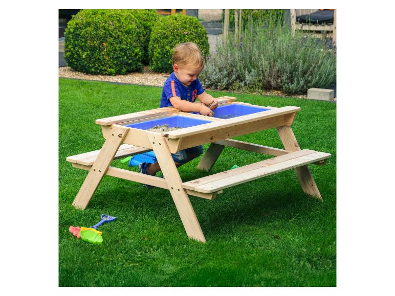 Kleinkind spielt mit Sand und Wasser an einem Holztisch mit blauen Behältern.