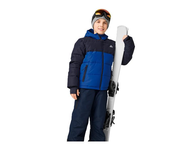Ein lächelnder Junge in blauer und schwarzer Skijacke, Hose und Skibrille, der Skier hält.