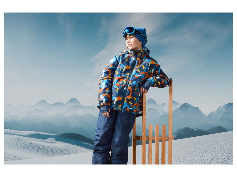 Ein Junge in einer Skijacke und mit Skibrille steht an einem Holzschlitten vor einer Bergkulisse.