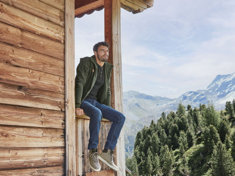 Mann in grünem Pullover und blauer Jeans sitzt auf einem Holzbalkon mit Blick auf die Berge.