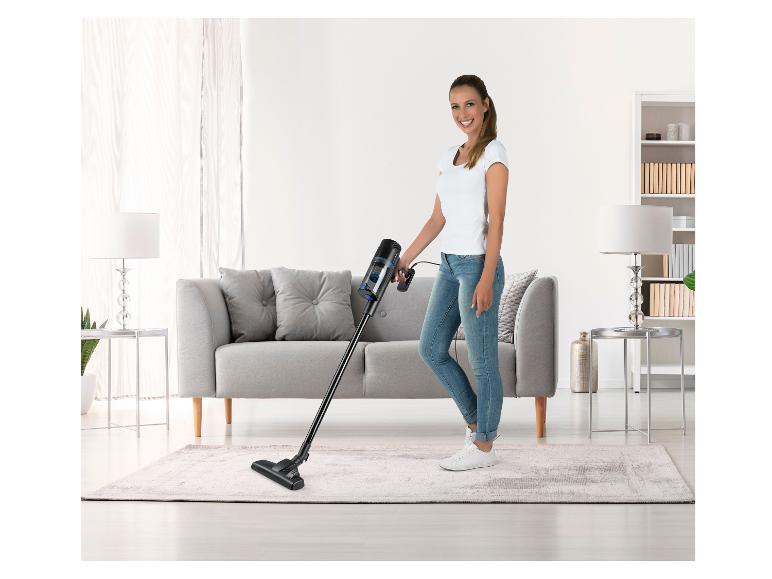 Lächelnde Frau saugt Teppich im Wohnzimmer mit einem Stielstaubsauger.