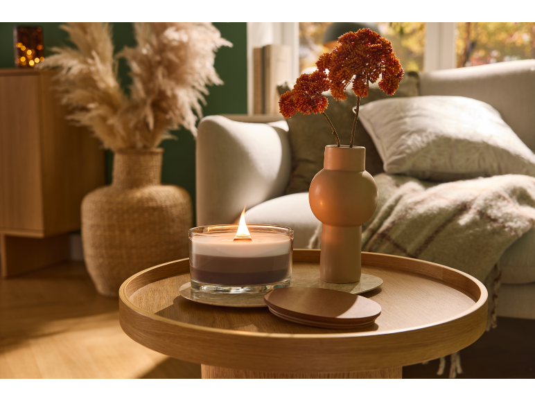 Brennende Kerze und Vase mit Herbstblumen auf einem Holztisch im Wohnzimmer.