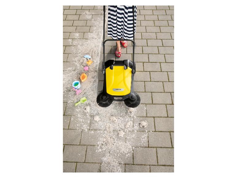 Frau in gestreiftem Kleid und roten Sandalen schiebt gelbe Kehrmaschine auf Pflaster, kehrt Sand.