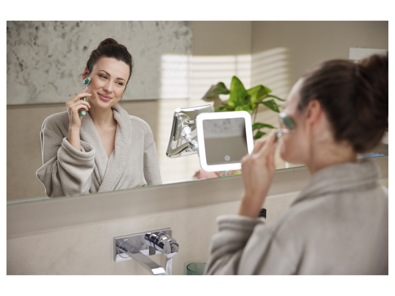 Eine Frau verwendet einen Jade-Roller und einen beleuchteten Spiegel im Badezimmer.