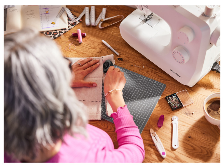 Eine Frau, die mit einem Rollmesser an Stoff arbeitet, mit einer Nähmaschine und anderem Nähzubehör daneben