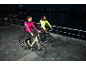 Zwei Personen in reflektierenden Fahrradjacken fahren nachts mit E-Bikes am Fluss entlang.