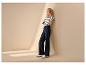 Frau in schwarz-weiß gestreiftem T-Shirt und dunkelblauer weiter Jeans, an eine beige Wand gelehnt.