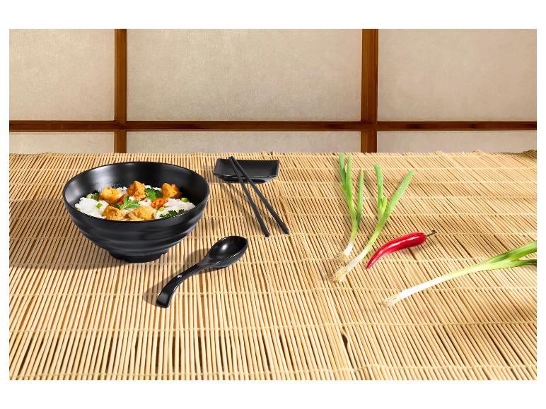 Schwarze Schale mit asiatischem Essen, Essstäbchen, Löffel, Frühlingszwiebeln und Chili auf einer Bambusmatte.