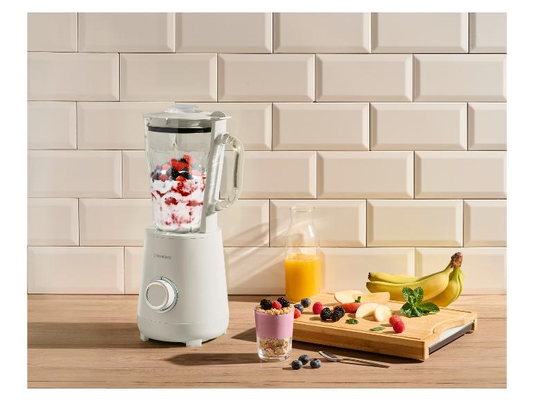 Ein Mixer gefüllt mit Beeren und Joghurt, daneben ein Glas Smoothie und frisches Obst.