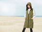 Eine Frau trägt eine lange, grüne, gesteppte Weste mit Kapuze am Strand.