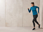 Eine Frau in einem blauen Sport-Oberteil und schwarzen Leggings rennt vor einer Betonwand.