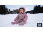 Lächelndes Baby in rosa Schneeanzug sitzt im Schnee, mit einem Haus und Bäumen im Hintergrund.