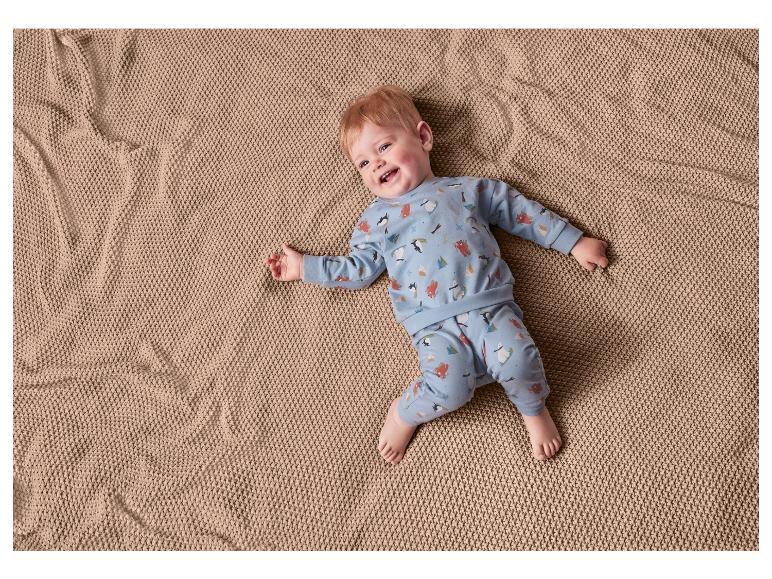 Baby in blauem Pyjama mit Pinguinen und Bären auf einer braunen Decke.