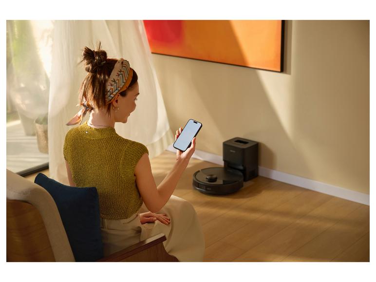 Frau steuert Saugroboter mit Smartphone im Wohnzimmer.