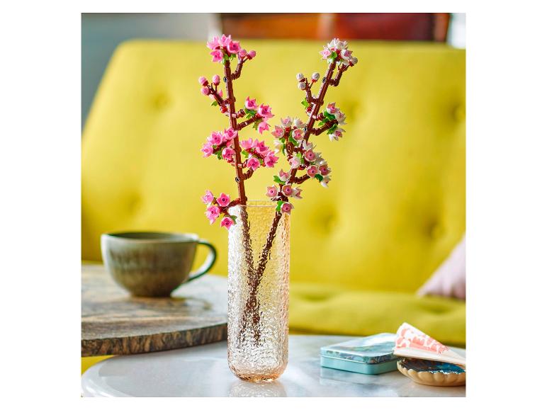 Vase mit Lego Botanicals Kirschblüten auf einem Tisch, mit einer Tasse und Dose im Hintergrund.