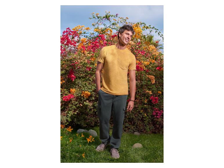 Mann in gelbem T-Shirt und dunkelgrüner Hose vor Blumen.