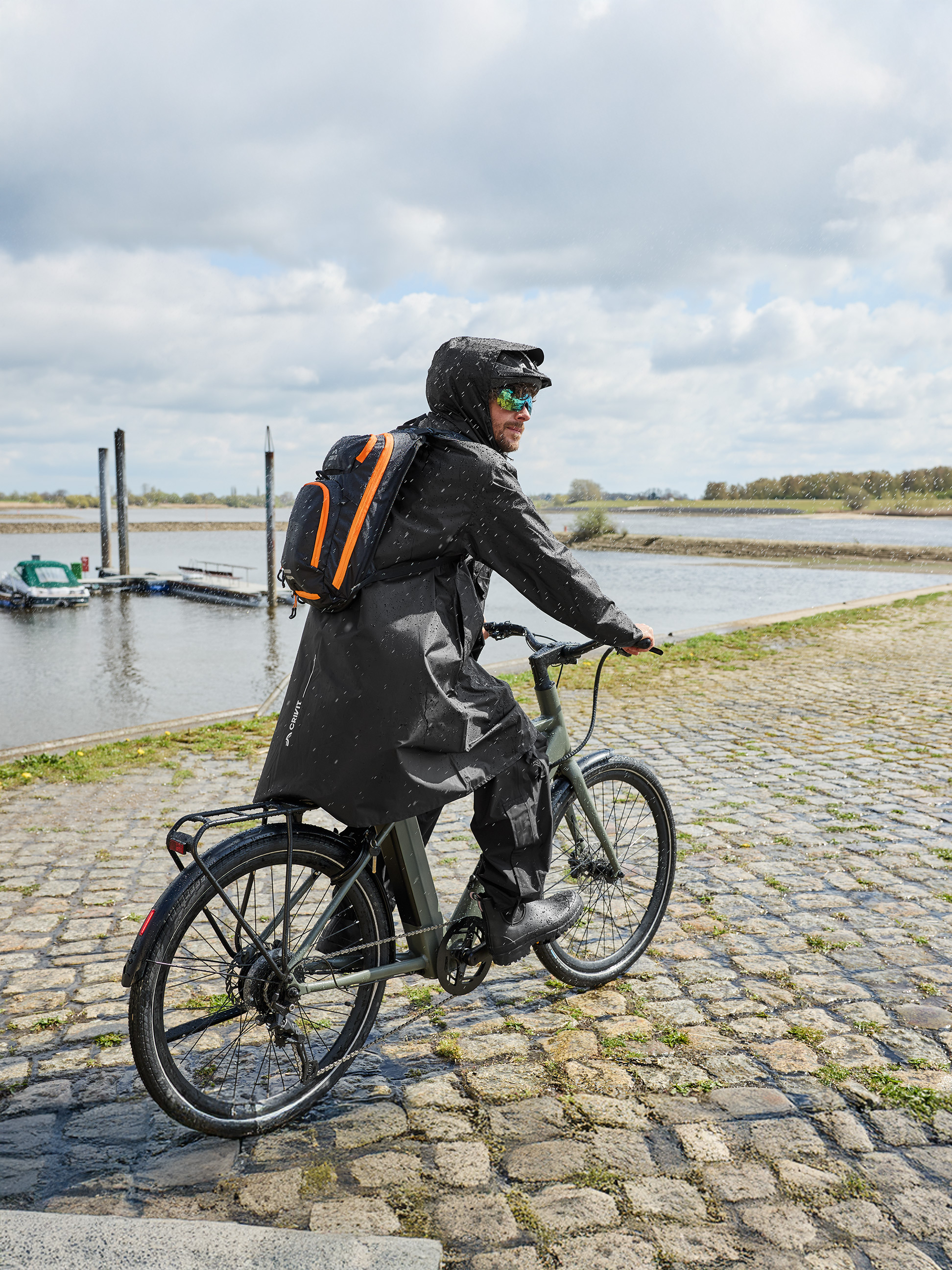 Mann in Regenjacke und Rucksack fährt bei Regen mit dem Fahrrad am Fluss.