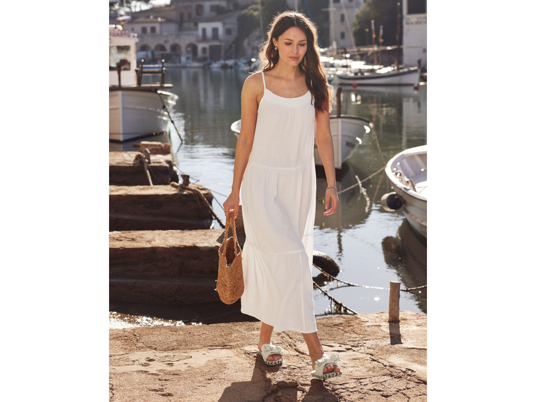 Eine Frau in einem weißen Kleid und Sandalen geht mit einer Korbtasche an einem Hafen entlang.