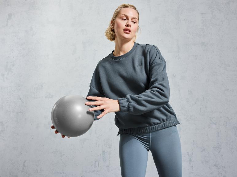 Frau in einem grauen Sweatshirt und Leggings hält einen Gymnastikball.