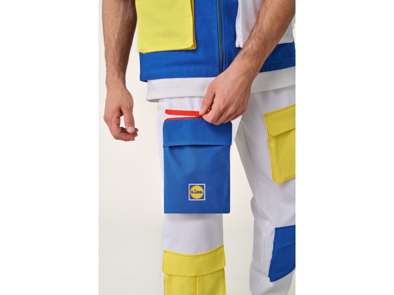 Detail eines Mannes in weißer Weste und Hose mit blauen und gelben Taschen, der eine Tasche mit rotem Reißverschluss öffnet.