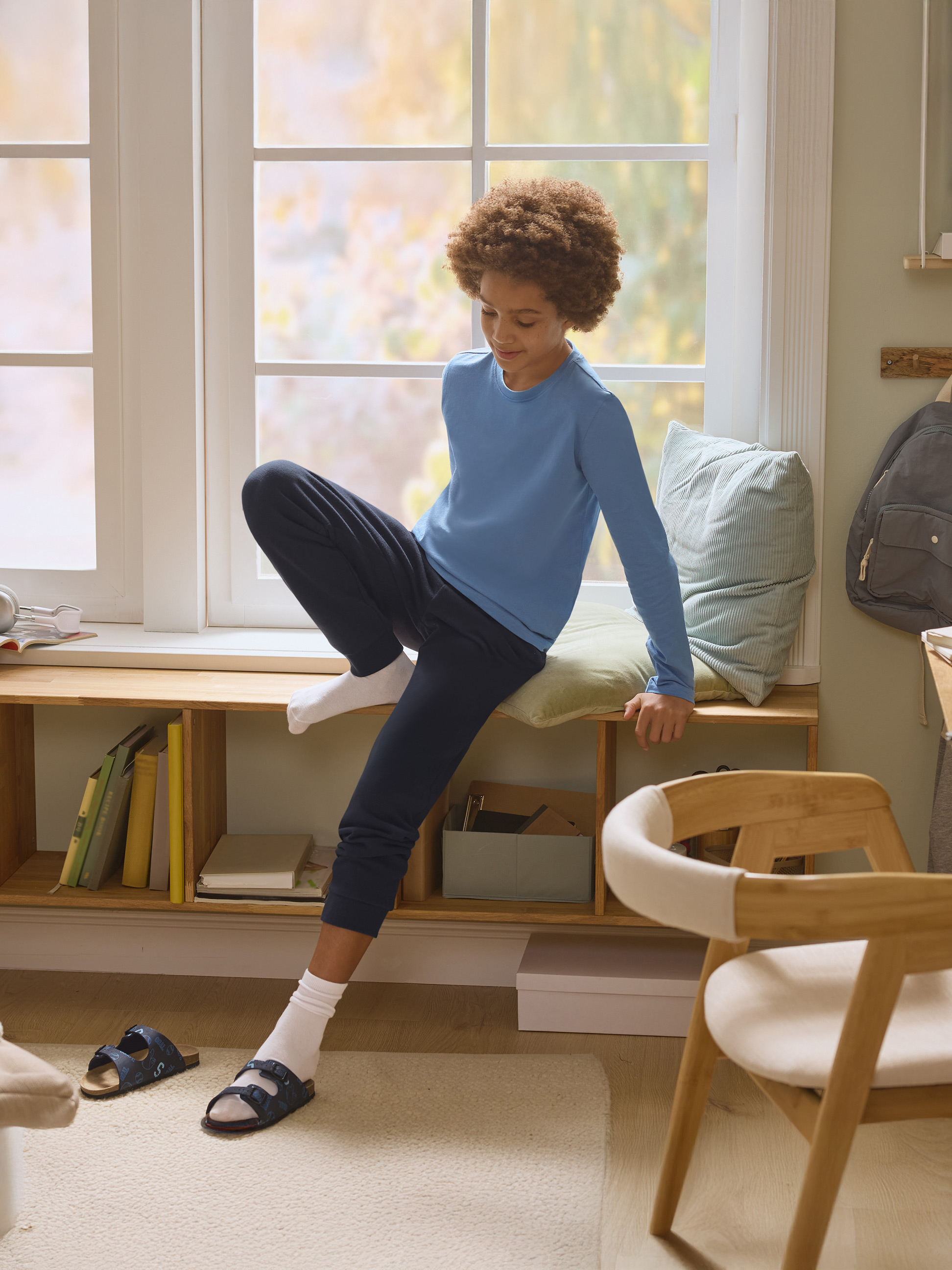 Junge im blauen Pyjama sitzt auf der Fensterbank mit Büchern und Kissen.