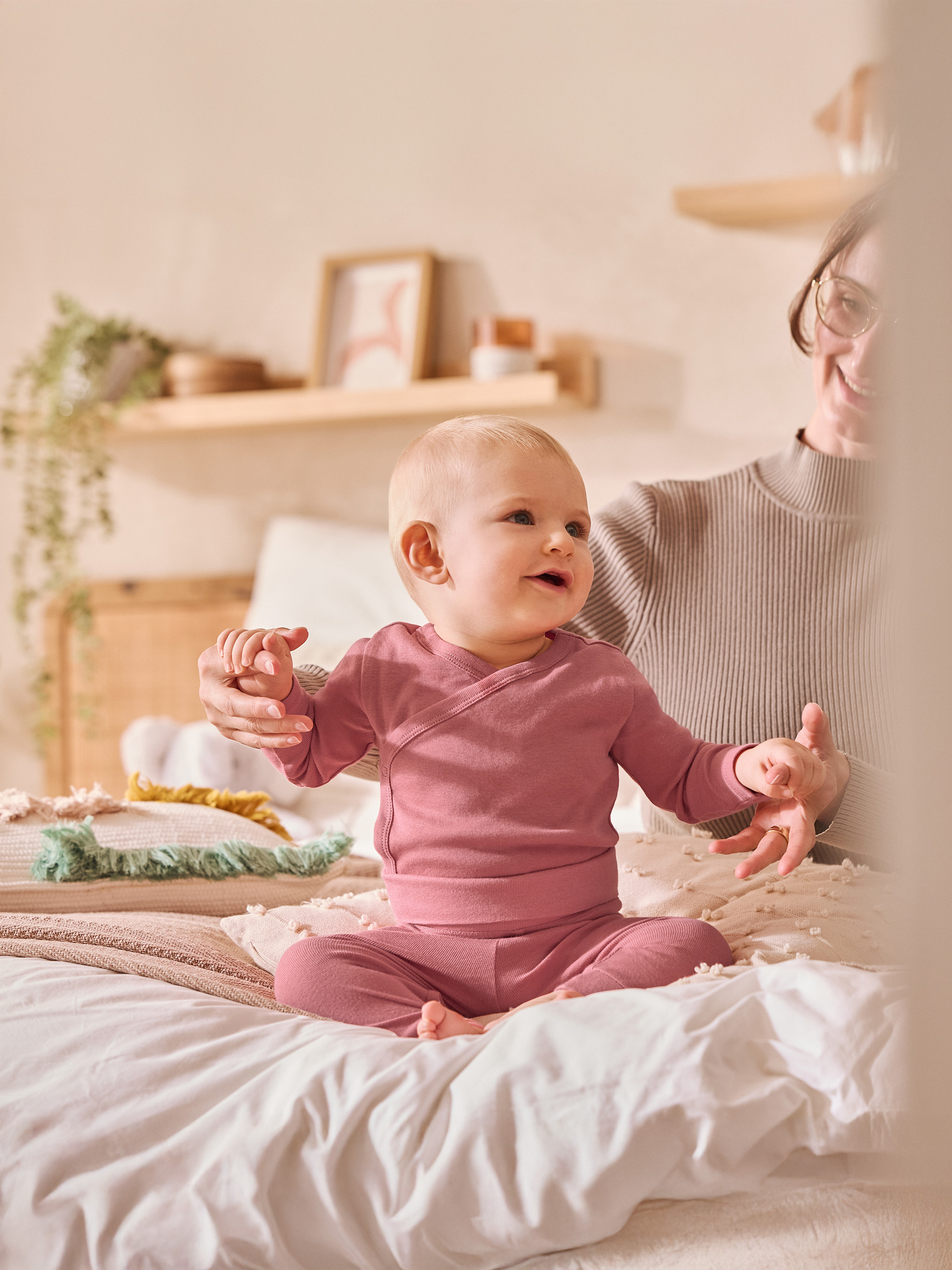Baby in rosa Strampler sitzt auf dem Bett und hält Händchen mit einem Erwachsenen.