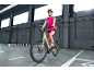 Lächelnde Frau in pinker Sportkleidung und Helm fährt ein graues E-Bike in einer Lagerhalle.