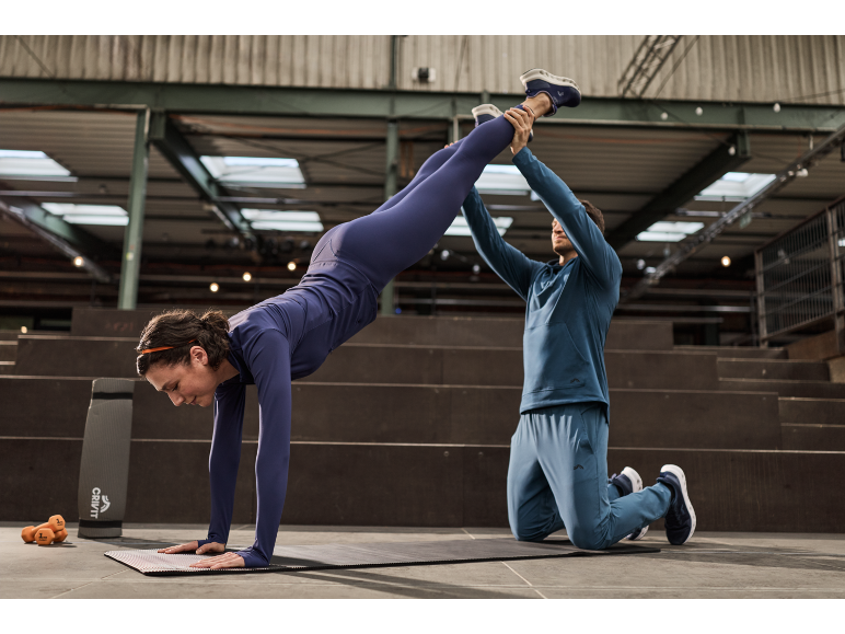Frau in blauem Sportoutfit macht einen Handstand auf einer Matte mit Hilfe eines Trainers.