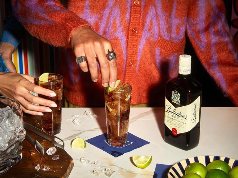 Zwei Gläser Getränk mit Eis und Limette, eine Flasche Whisky und eine Schale Limetten auf einem Tisch.