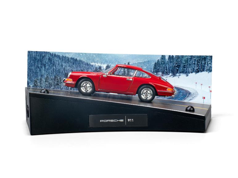 Rotes Porsche 911 Modellauto auf schwarzem Sockel mit Winterlandschaft im Hintergrund.