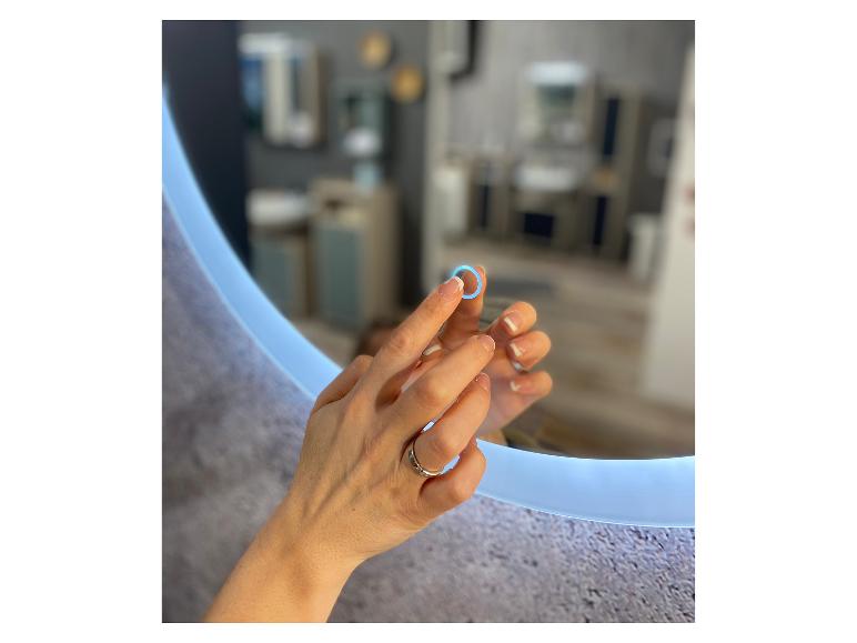 Frauenhände berühren einen runden beleuchteten Spiegel, der ein modernes Badezimmer reflektiert.