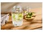 Getränkespender aus Glas mit geschnittenen Limetten, Gläsern und ganzen Limetten auf einem Holztisch.