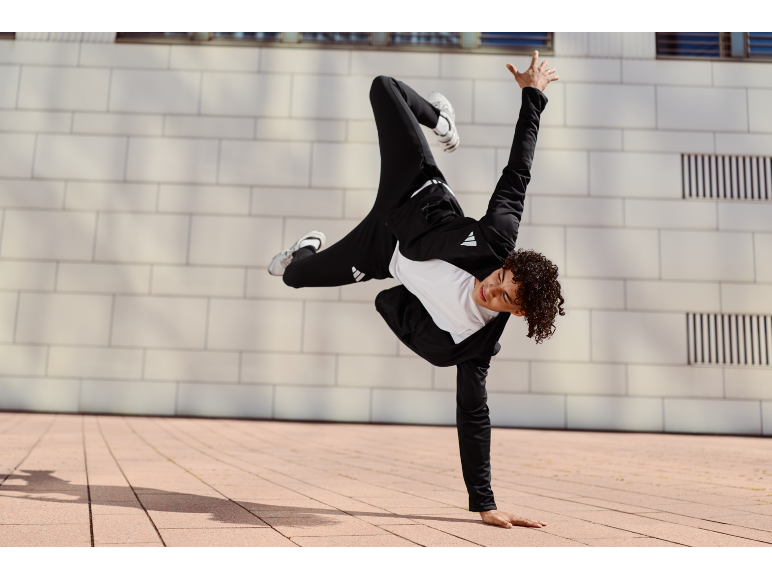 Junger Mann in schwarzem Trainingsanzug und weißem T-Shirt beim Breakdance im Freien.