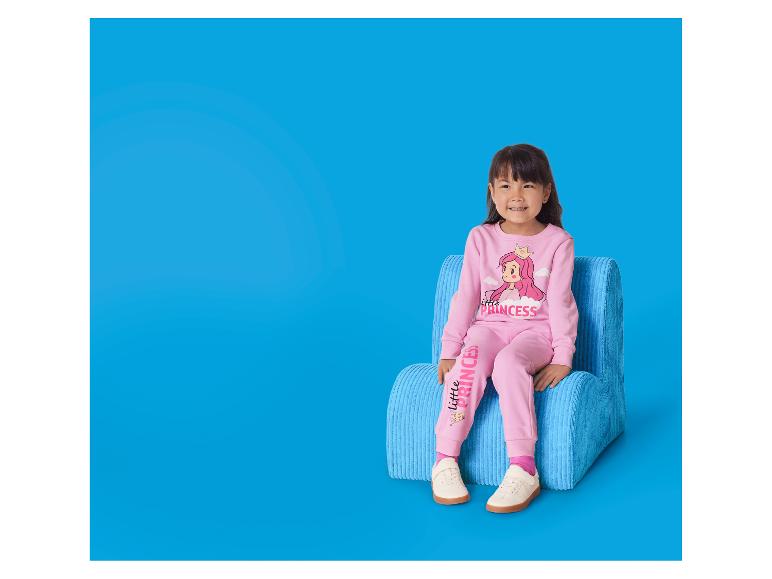 Kleines Mädchen in rosa Pyjama mit Prinzessinnenmotiv sitzt auf einem blauen Stuhl.