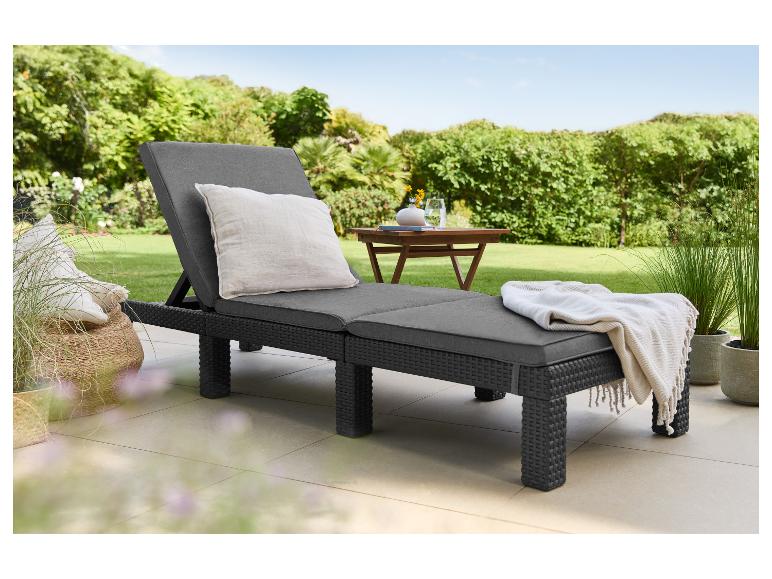 Dunkelgraue Sonnenliege mit Kissen und Decke auf einer Terrasse mit Garten im Hintergrund.