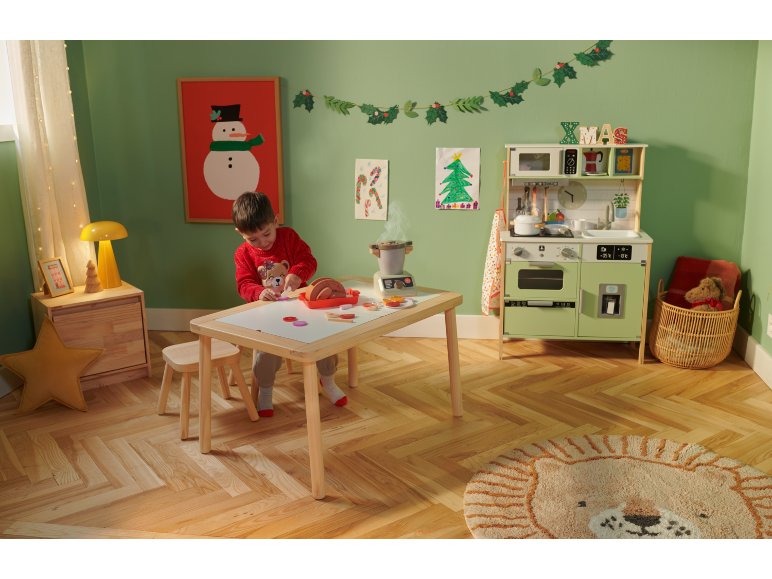 Kind spielt mit einer Holzküche und Spielzeugessen in einem weihnachtlich dekorierten Zimmer.
