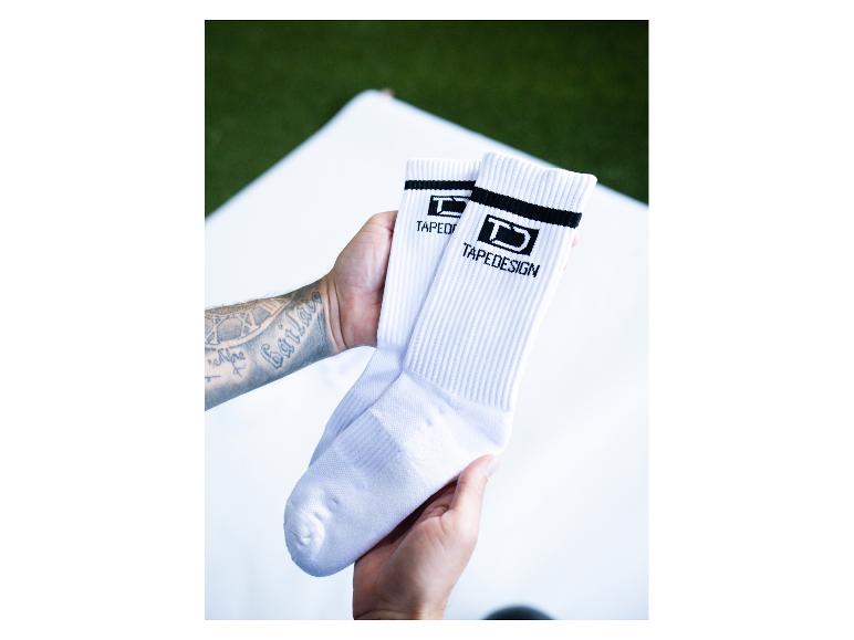 Weiße Tapedesign-Socken mit schwarzen Streifen, gehalten von tätowierten Händen.