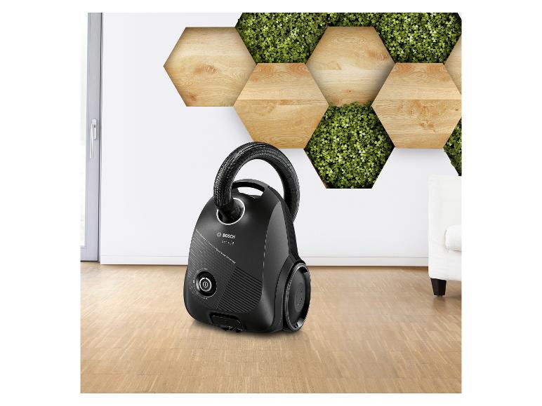 Schwarzer Staubsauger auf Holzboden vor einer dekorativen Wand aus Holz und Pflanzen.