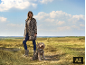 Frau in Leopardenmantel und passender Hund auf einem Feld.