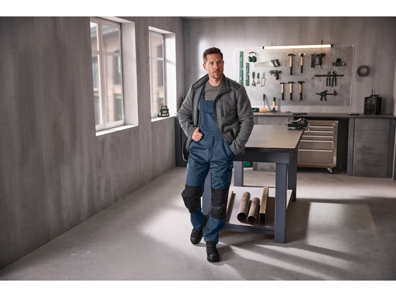 Mann in blauer Latzhose und grauer Fleecejacke in einer Werkstatt