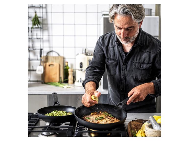 Ein Mann kocht Fisch und Gemüse in zwei Pfannen auf einem Gasherd.