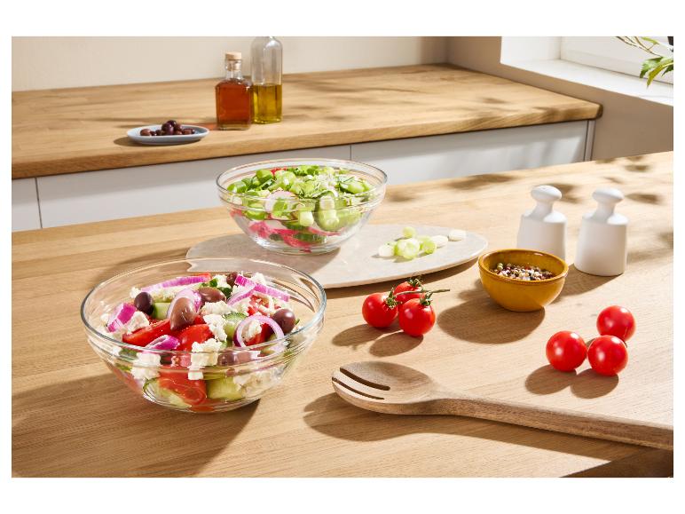 Frische Salate in Glasschalen auf einer Holztheke mit Dressings und Tomaten.