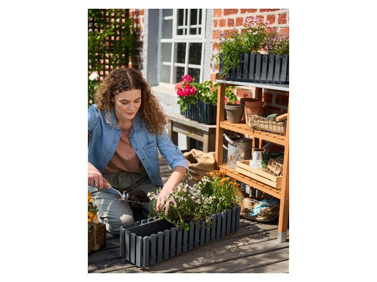 Frau pflanzt Blumen in einem grauen Pflanzgefäß auf einer Holzterrasse, mit Gartenregal.