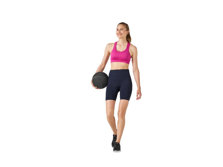 Frau in pinkem Sport-BH und dunkelblauen Shorts, hält einen schwarzen Medizinball.
