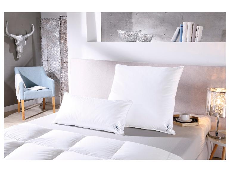 Zwei weiße Kissen mit Häussling-Logo auf einem Bett mit Bettdecke, neben Stuhl und Deko.